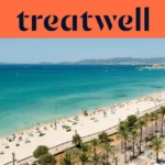 Vista aérea da Playa de Palma, em Maiorca, com uma longa curva de areia dourada, filas de chapéus de sol, água turquesa e calma e uma avenida marginal ladeada de palmeiras junto à cidade, com colinas ao fundo sob um céu azul e limpo.