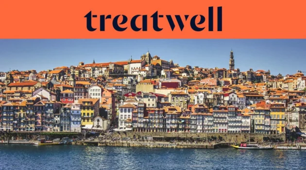 O passeio ribeirinho da Ribeira, no Porto, com filas de casas coloridas e telhados de telha a subir a encosta a partir do rio Douro, e a Torre dos Clérigos no horizonte sob um céu azul limpo.