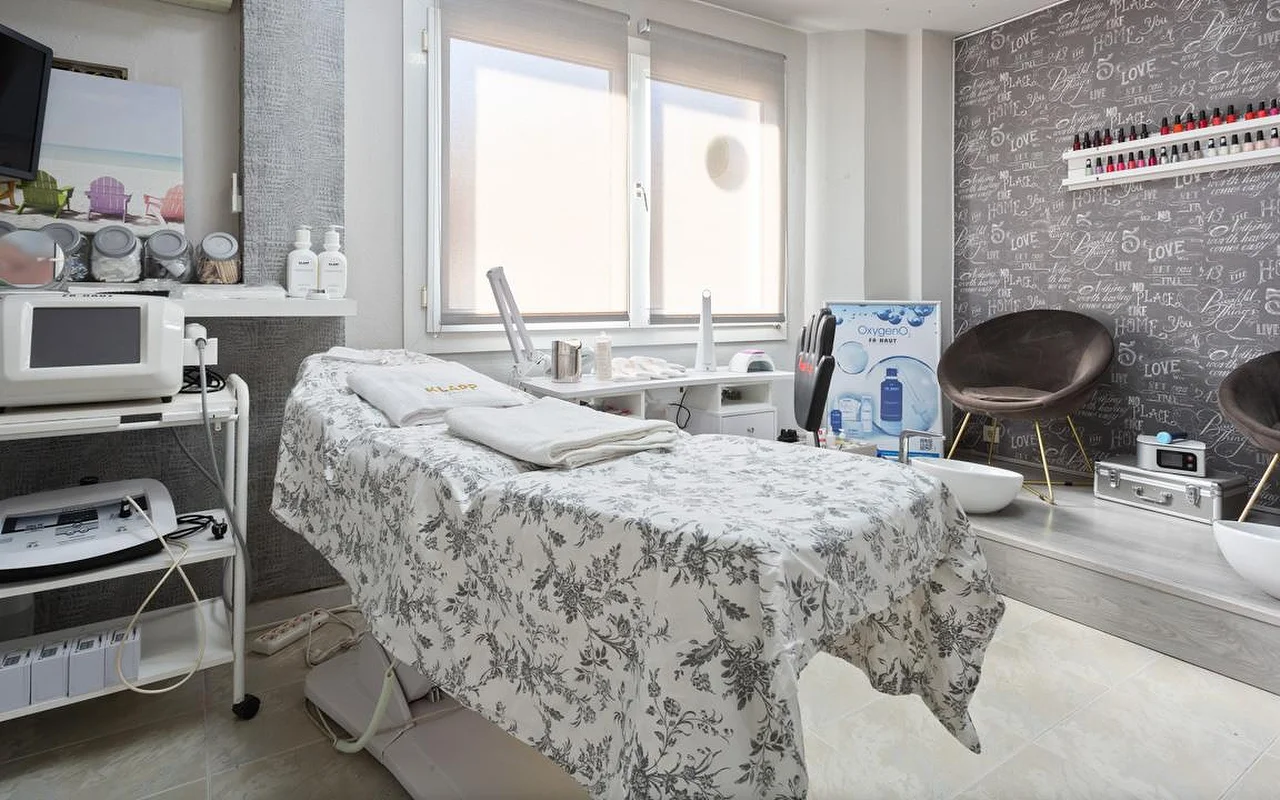 Uma sala de tratamentos de beleza luminosa, com uma marquesa vestida com roupa de linho de padrão floral cinzento e toalhas brancas dobradas, aparelhos de estética em carrinhos à esquerda, uma janela com o estore meio descido e uma zona elevada com uma cadeira de veludo castanho e bacias de pedicure junto a uma parede decorativa com letras manuscritas.