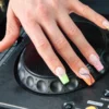 Uma mão com a electric mani pousada no jog wheel de um leitor de DJ Pioneer, com french tips em néon de padrão animal e em espiral