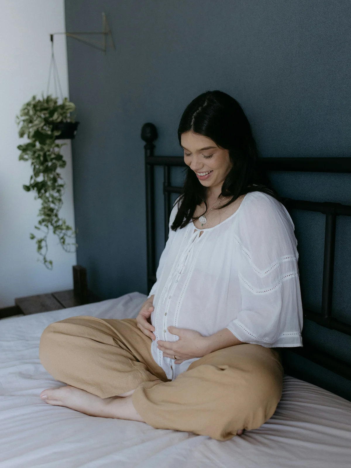 Uma mulher grávida sentada de pernas cruzadas numa cama, de camisa branca e calças bege, com as mãos sobre a barriga e um sorriso virado para baixo