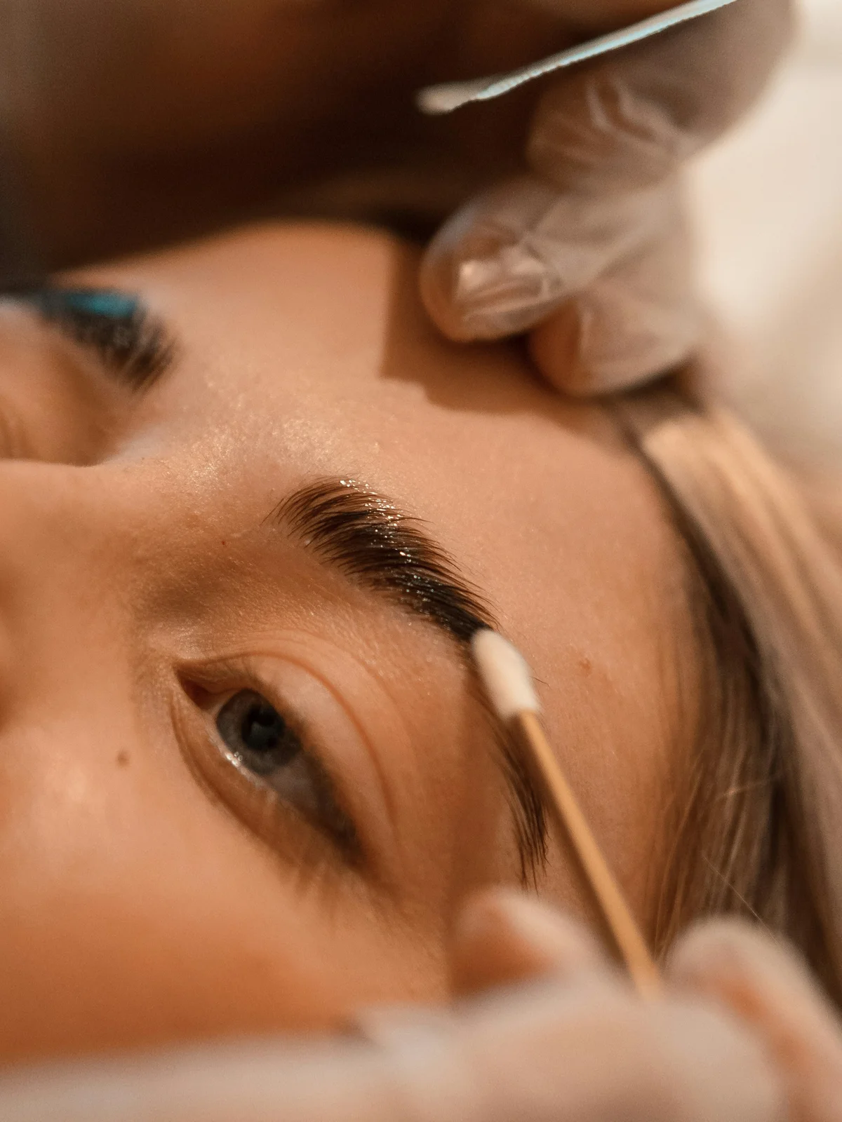 Grande plano de uma cliente deitada enquanto uma esteticista faz uma laminação de sobrancelhas