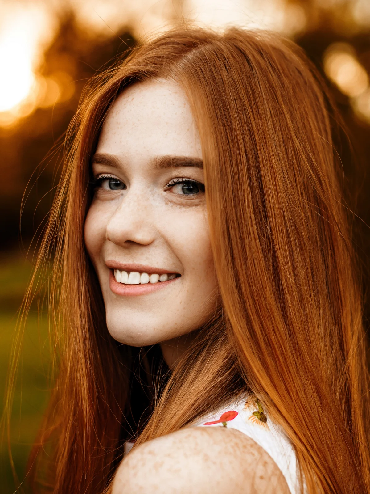 Uma mulher de cabelo comprido e ruivo, com as sobrancelhas arranjadas e definidas, sorri enquanto passeia ao ar livre na natureza com a luz do fim de tarde