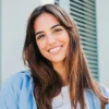Uma mulher de cabelo comprido e escuro, com as sobrancelhas bem definidas, sorri ao ar livre em frente a um edifício com portadas verdes