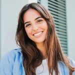 Uma mulher de cabelo comprido e escuro, com as sobrancelhas bem definidas, sorri ao ar livre em frente a um edifício com portadas verdes