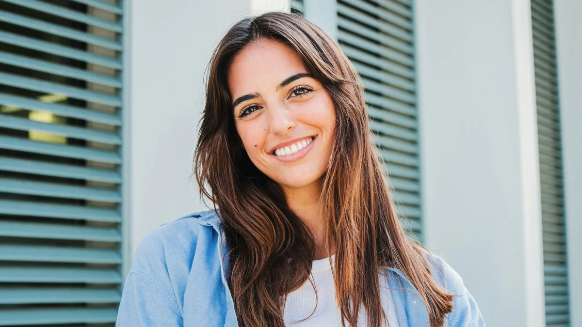 Uma mulher de cabelo comprido e escuro, com as sobrancelhas bem definidas, sorri ao ar livre em frente a um edifício com portadas verdes