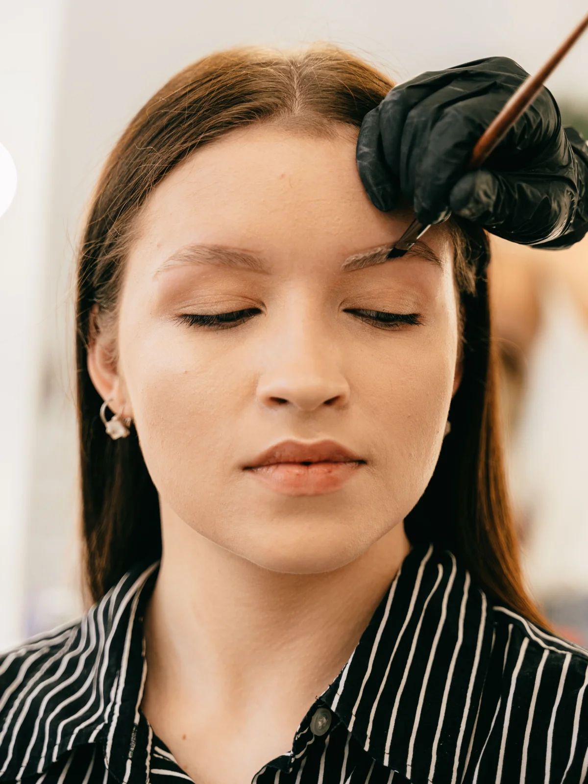 Uma cliente sentada no salão de olhos fechados enquanto uma esteticista aplica com cuidado a coloração com um pincel fino