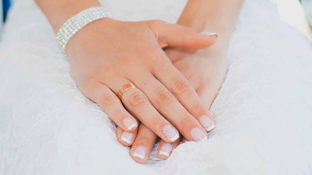 As mãos de uma mulher repousam no colo; usa um vestido de noiva em renda e uma manicure francesa cuidada numa combinação clássica de nude e branco em unhas curtas quadradas-ovais, com um delicado adorno nos dedos anelares