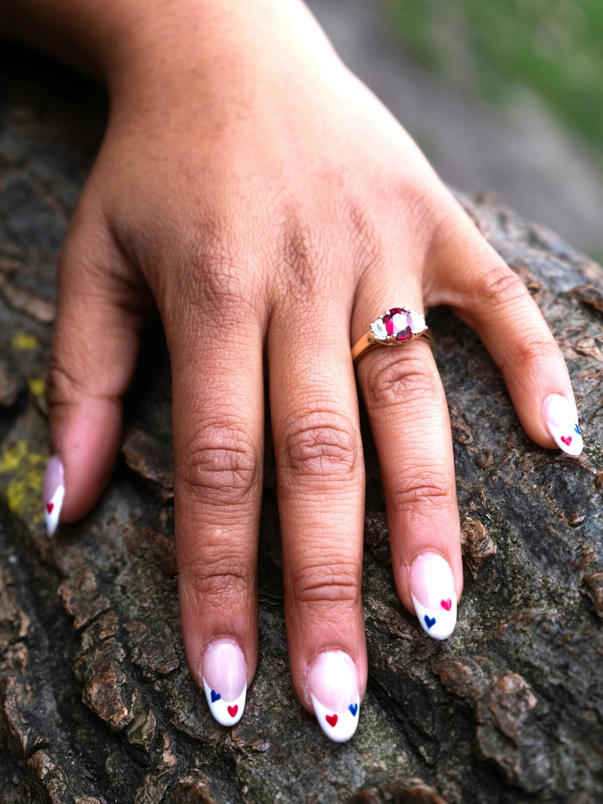 A mão de uma mulher pousada sobre um ramo mostra uma manicure francesa em unhas ovais, decoradas com pequenos corações vermelhos e azuis