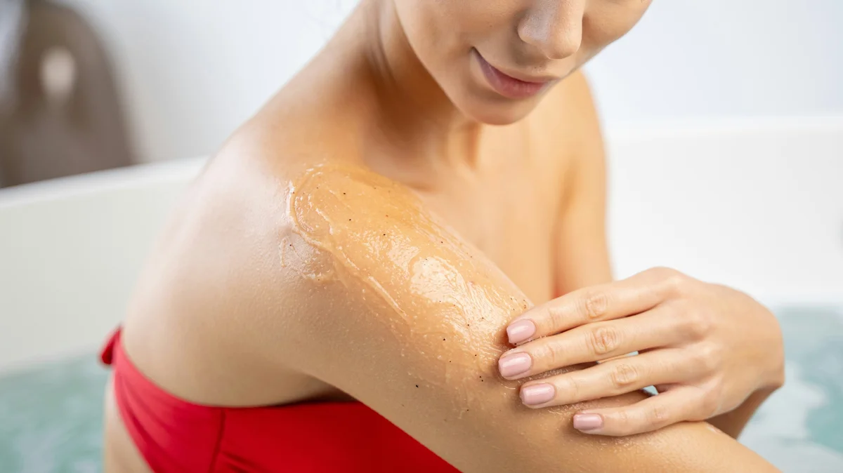 Grande plano de uma mulher de biquíni vermelho a aplicar um esfoliante na parte superior do braço numa casa de banho luminosa e arrumada