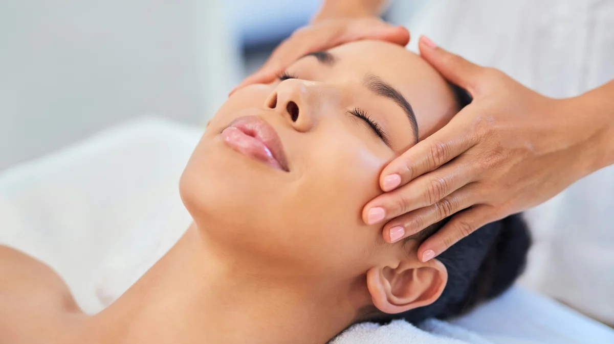 Uma mulher deitada num centro de estética enquanto uma esteticista lhe pousa suavemente as mãos na cabeça durante um tratamento facial