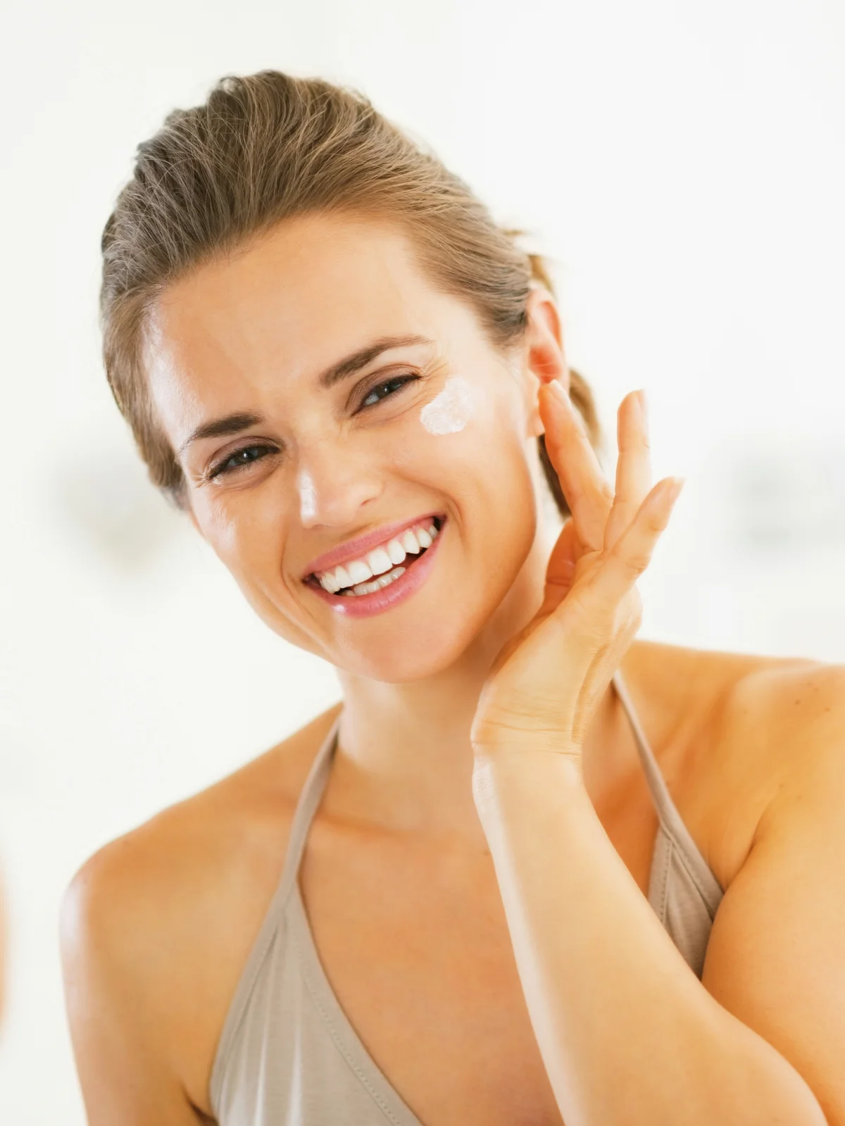 Grande plano de uma mulher sorridente com o cabelo apanhado a aplicar protetor solar no rosto