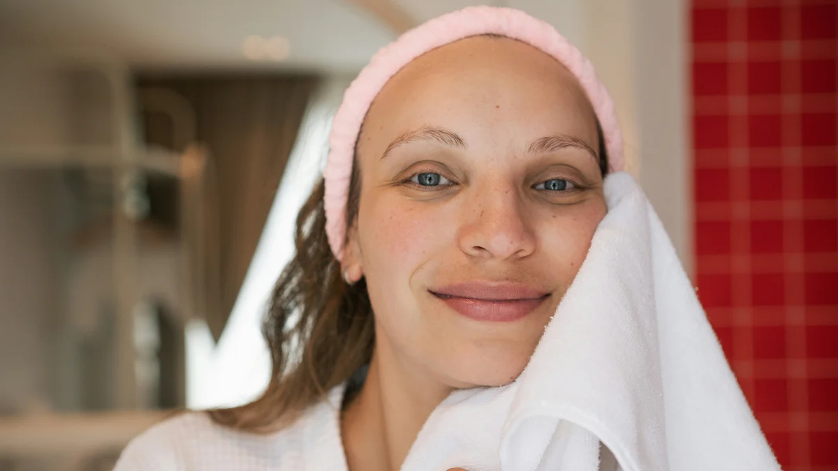 Uma mulher de pele limpa e luminosa sorri para a câmara com uma bandolete cor-de-rosa enquanto seca o rosto acabado de limpar com uma toalha branca