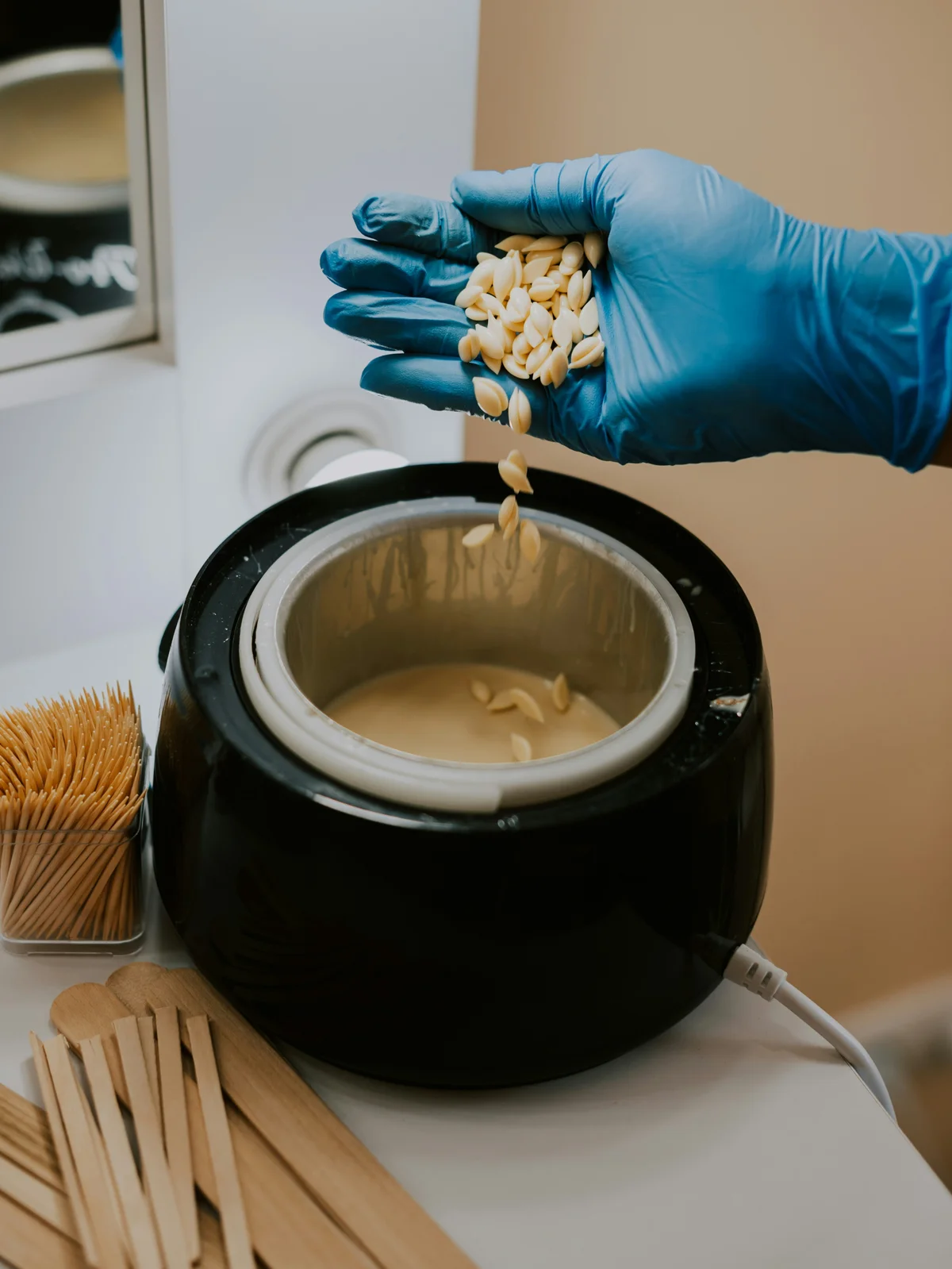 Grande plano da mão enluvada de uma esteticista a misturar pérolas de cera quente num recipiente, pronta para a marcação na cabine
