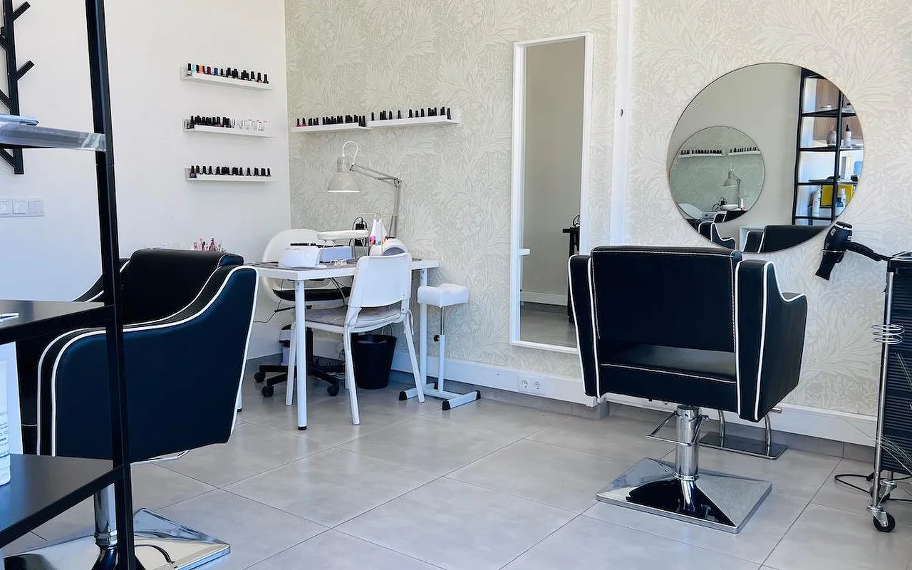 Um compacto salão de cabelo e unhas com cadeiras de styling pretas debruadas a branco, um espelho redondo e um espelho alto sobre um papel de parede cinzento de flores, e prateleiras de parede alinhadas de vernizes.