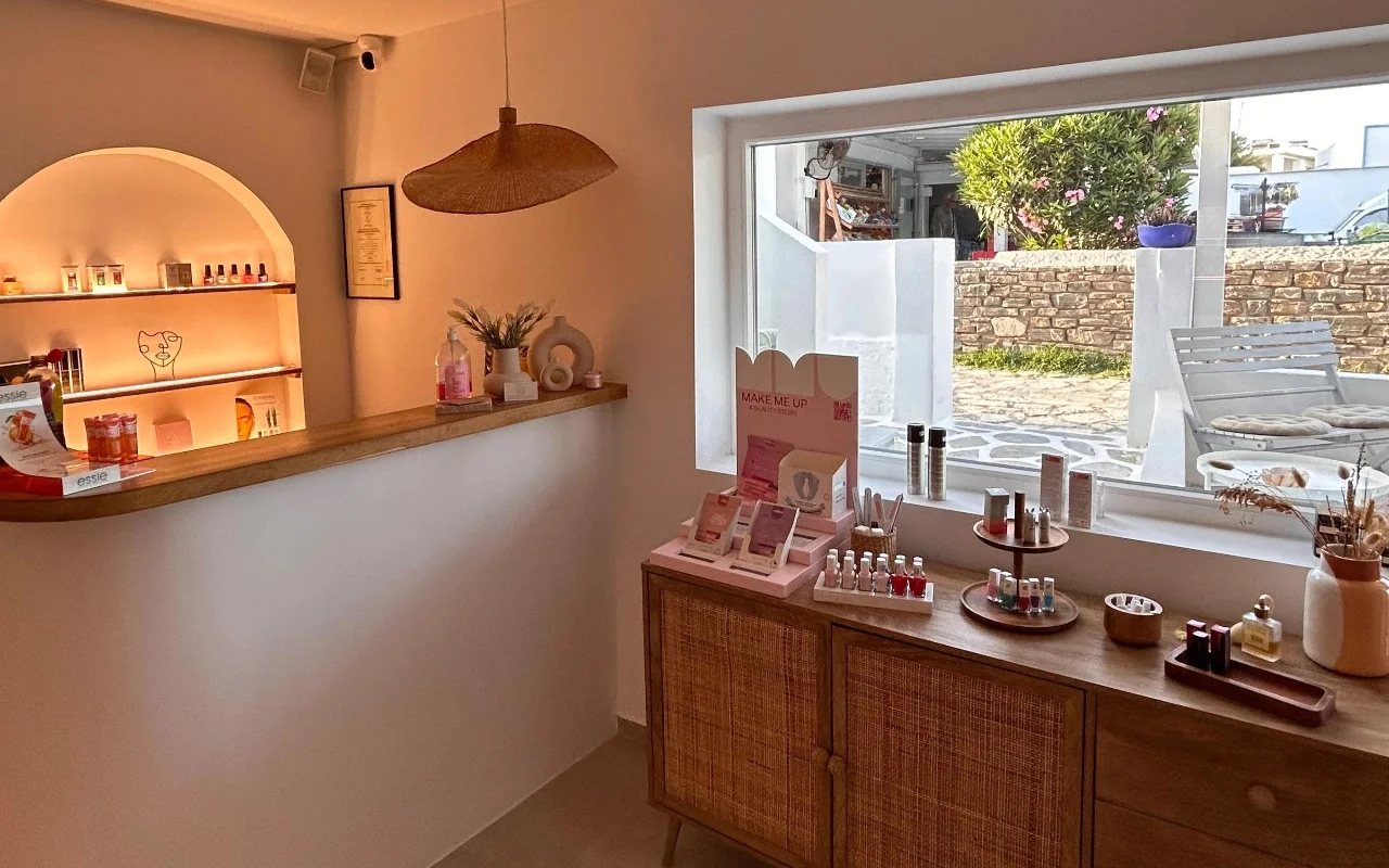 O interior do BeautiQ Esthetics and More em Paros, com um nicho em arco suavemente iluminado cheio de vernizes, um candeeiro suspenso de rotim entrançado e uma grande janela com vista para um muro de pedra e um jardim soalheiro.