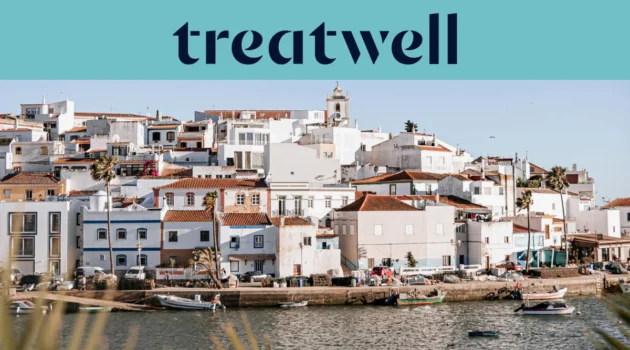 Uma aldeia piscatória algarvia caiada de branco, com casas de telhados de terracota a subir por uma encosta acima de um plano de água calmo, pequenos barcos de pesca atracados ao longo do cais, palmeiras e a torre de uma igreja sob um céu azul límpido.