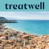 Vista ampla sobre uma cidade costeira siciliana, com edifícios antigos de telhados em terracota, uma meia-lua de praia de areia, um mar turquesa cristalino e um promontório rochoso que se ergue de um dos lados.