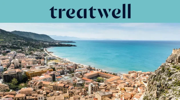 Vista ampla sobre uma cidade costeira siciliana, com edifícios antigos de telhados em terracota, uma meia-lua de praia de areia, um mar turquesa cristalino e um promontório rochoso que se ergue de um dos lados.