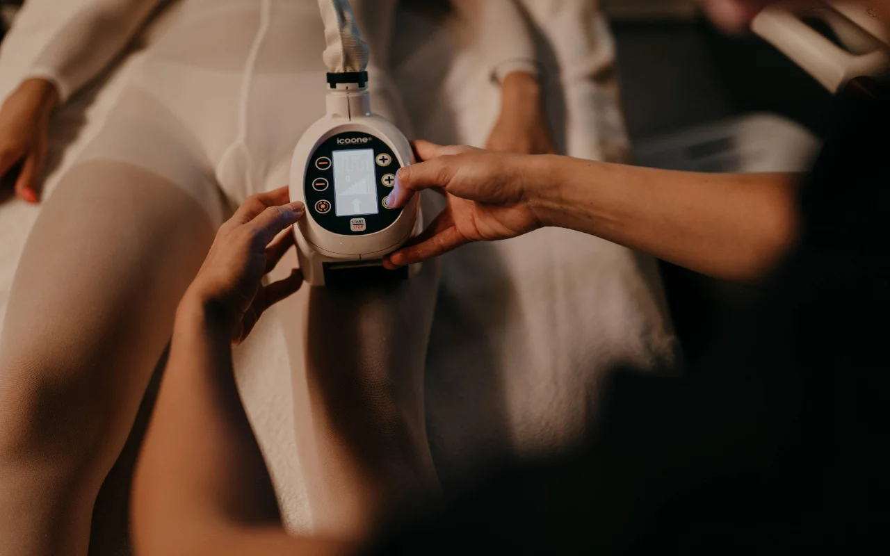 Um tratamento de remodelação corporal na Beauty Room Romina Bongiovanni, com uma profissional a aplicar um aparelho icoone na perna de uma cliente com fato branco de tratamento, numa luz quente e suave.