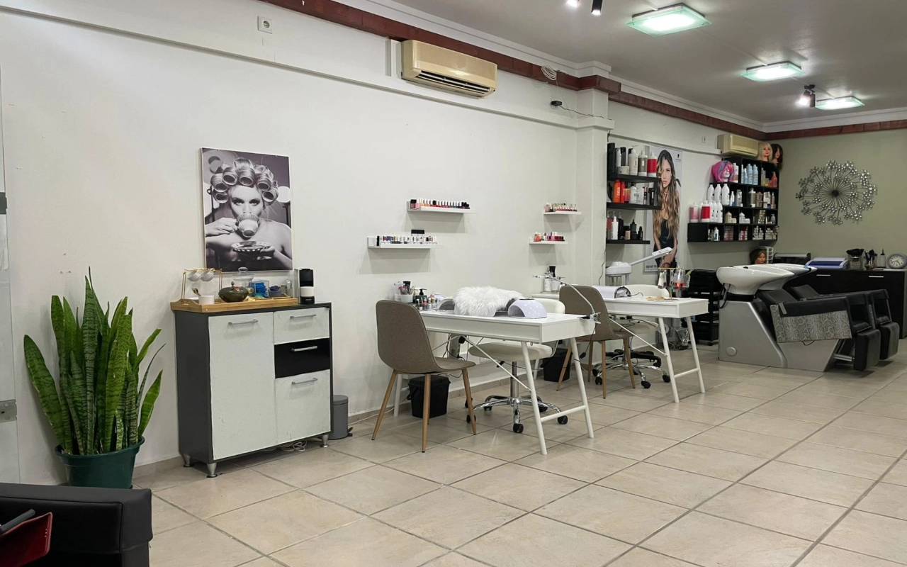 Um salão de cabelo e beleza com duas mesas de manicure brancas e cadeiras cor de toupeira, um lava-cabeças e prateleiras de produtos ao longo da parede, uma alta espada-de-são-jorge a um canto e uma grande fotografia a preto e branco de uma mulher de rolos no cabelo.