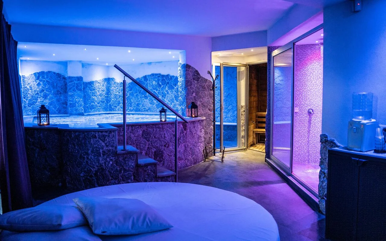 Uma suite de spa privada na Bellezza e Benessere, em Taormina, iluminada de azul e roxo, com uma banheira de hidromassagem encastrada num revestimento de pedra, uma sauna de madeira e, ao fundo, um banho turco em mosaico, e uma cama redonda em primeiro plano.