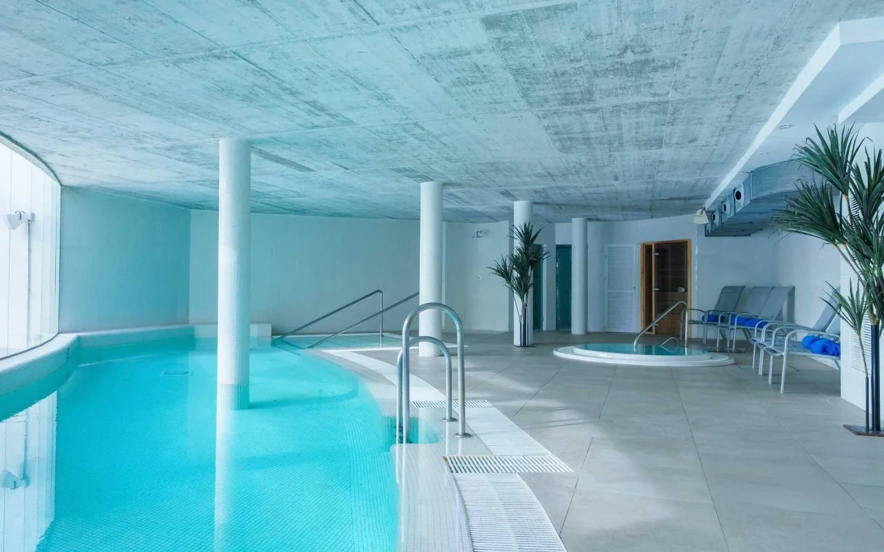 A piscina interior de spa da Benalmádena Beach, com um tanque turquesa curvo, um teto de betão à vista, um pequeno jacuzzi e espreguiçadeiras entre altas palmeiras em vaso.
