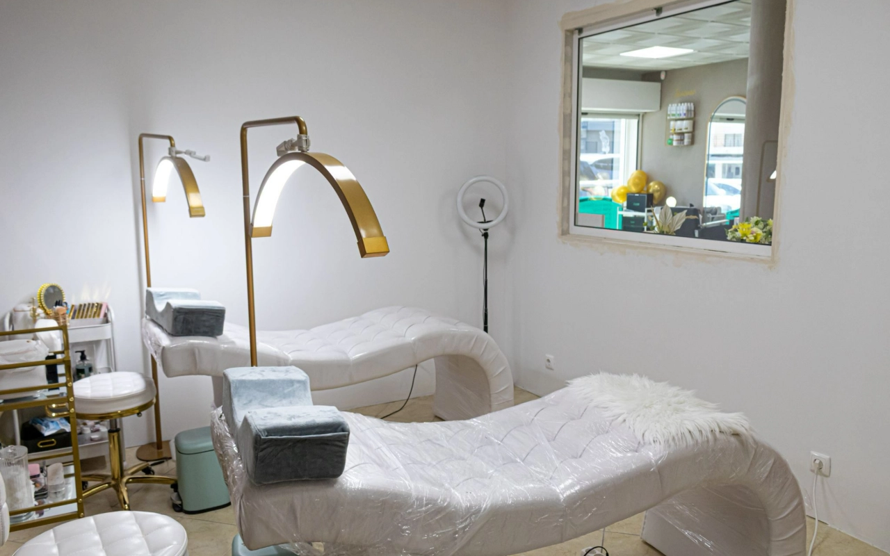 Uma luminosa sala de tratamentos de beleza com duas marquesas brancas acolchoadas, altos candeeiros de arco dourados e um aro de luz, com uma janela interior virada para a receção.