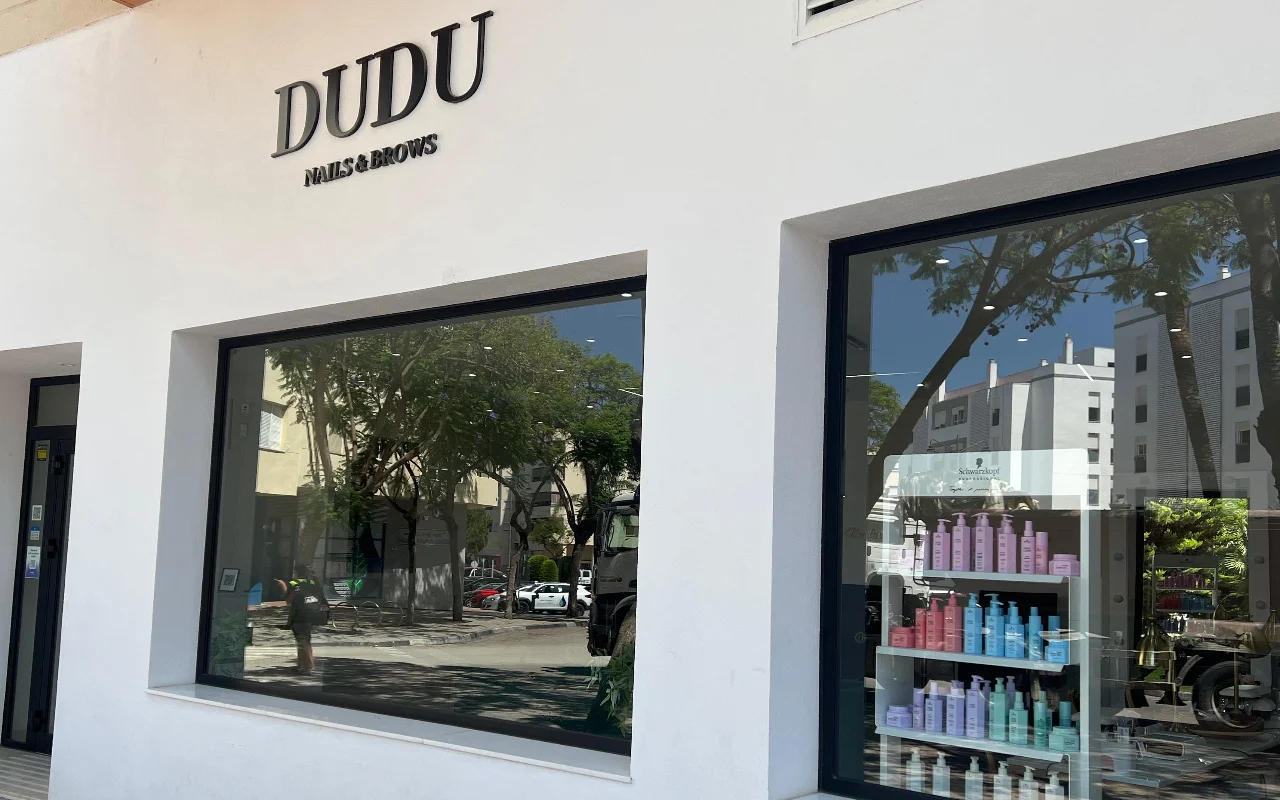 A montra branca da Dudu Nails and Brows em Estepona, com o nome em letras escuras sobre amplas vitrinas que mostram filas de produtos para o cabelo.