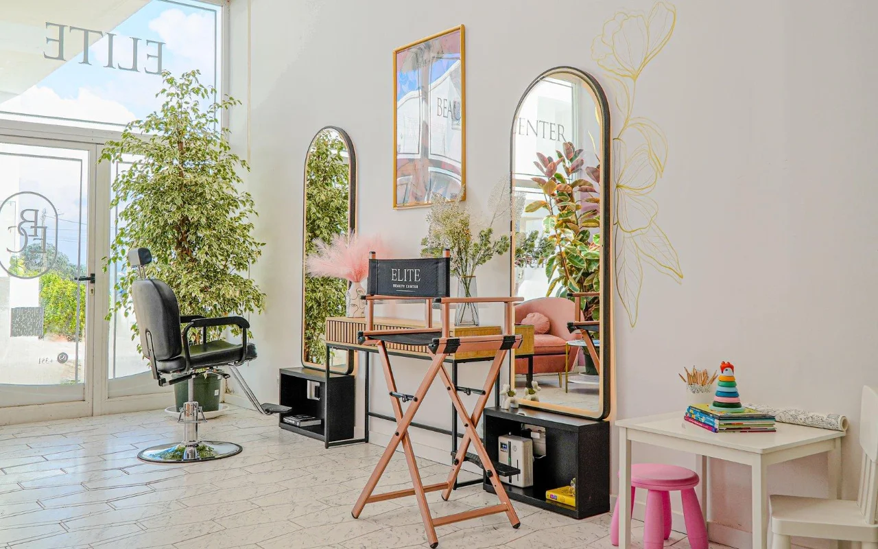 Um salão de beleza luminoso e arejado, todo branco, com espelhos em arco de moldura dourada, uma cadeira de cabeleireiro preta e uma cadeira dobrável de realizador, altas plantas verdes, apontamentos de rosa e um pequeno cantinho para crianças junto à janela.