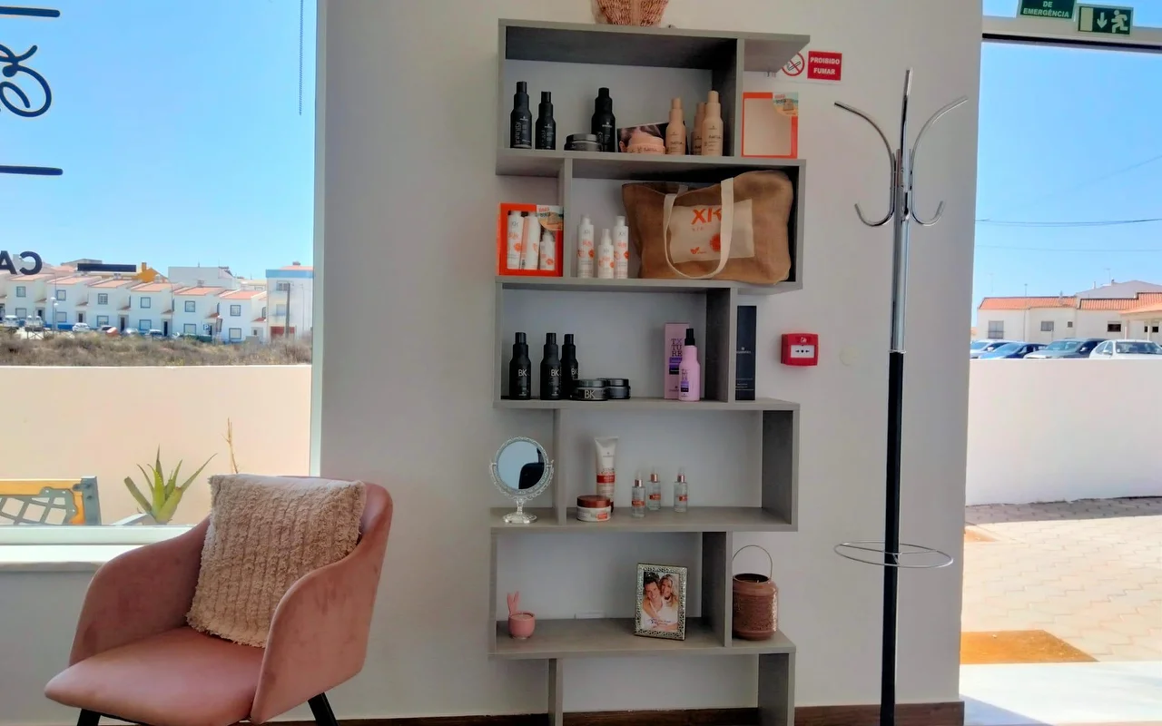 Um canto de salão junto a uma grande janela, com uma prateleira cinzenta em degraus cheia de produtos de cabelo e uma fotografia emoldurada, um cadeirão de veludo rosa-velho com uma almofada, e vista para casas baixas caiadas de branco sob um céu azul.