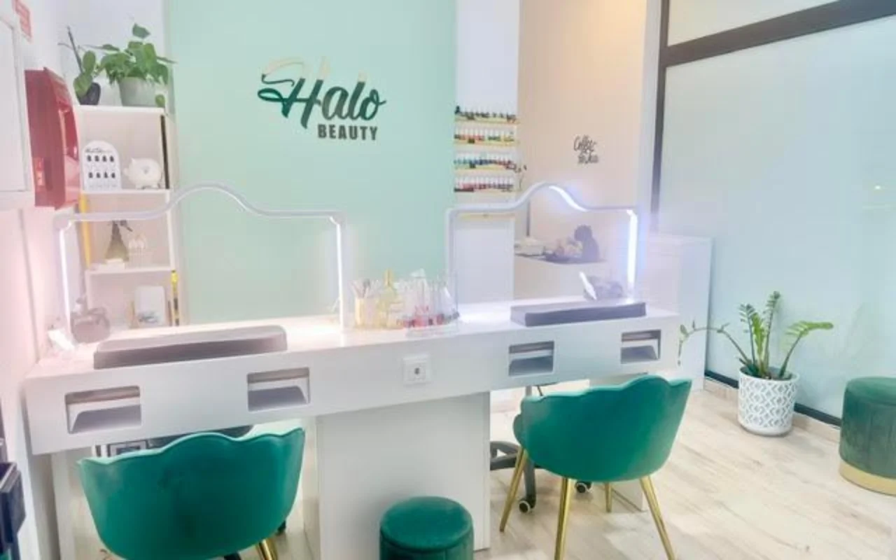 Um salão de unhas com uma parede de destaque verde-menta com o logótipo da Halo Beauty, uma dupla bancada de manicure branca com divisórias de acrílico iluminadas, dois cadeirões em veludo verde-esmeralda, um expositor de vernizes na parede e plantas em vaso.
