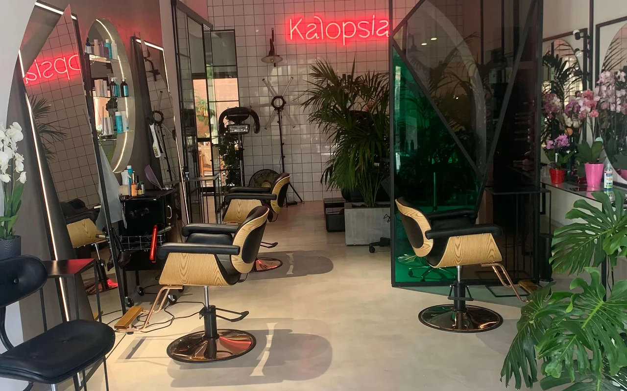 O espaço de styling do salão Kalopsia em Ermoupoli, Syros, com cadeiras em madeira clara e preto, espelhos em arco retroiluminados, plantas frondosas e um letreiro néon rosa com a palavra Kalopsia.