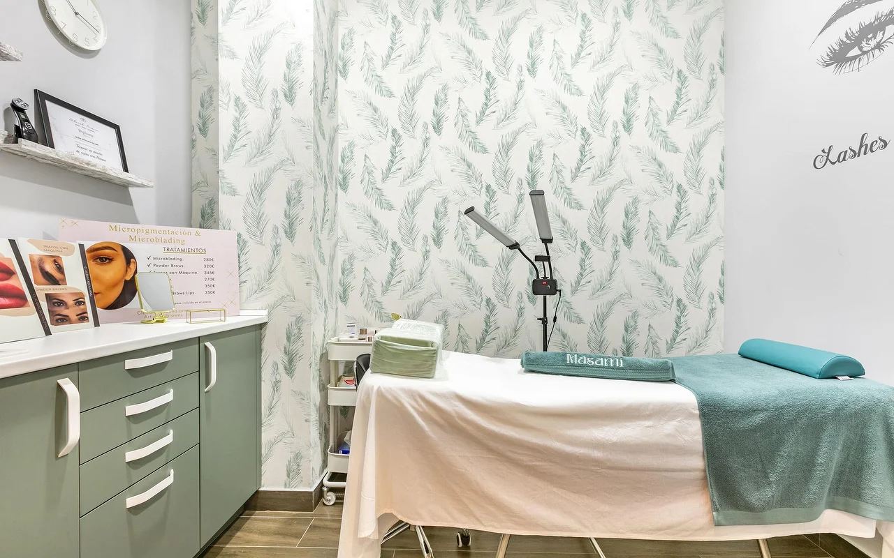 Uma cabine de tratamento na Masami Belleza Natural em Fuengirola, com uma marquesa, um papel de parede de penas, um candeeiro-lupa e uma tabela de preços de micropigmentação.