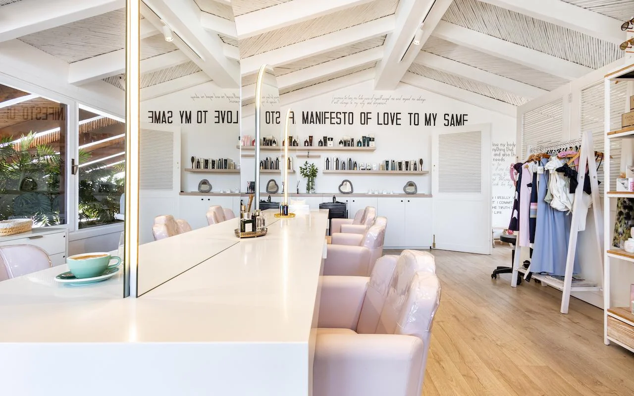 O interior luminoso da Milk & Roses em Puente Romano, Marbella, com um balcão de styling branco, cadeiras rosa-claro, um teto de vigas e prateleiras de produtos ao longo da parede.