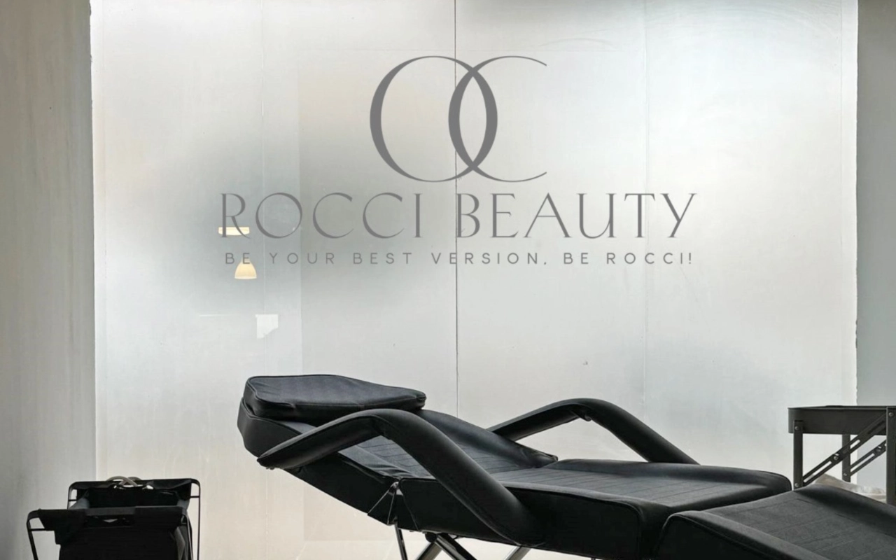 Uma sala de tratamentos com uma marquesa reclinável preta e um pequeno carrinho de equipamento, diante de uma parede de vidro fosco com o logótipo e o lema da Rocci Beauty.