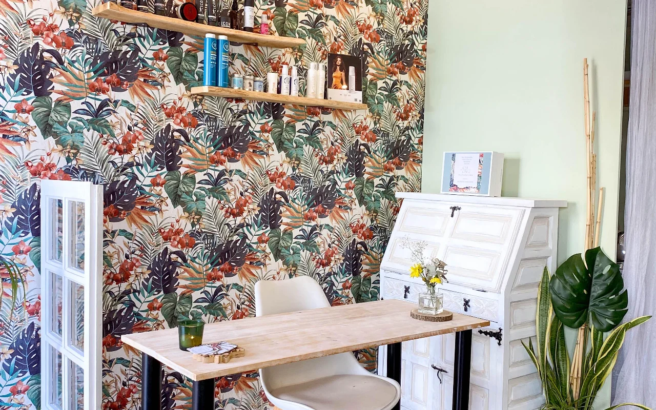 Um posto de styling na Shangara Lora Organics em Marbella, com um papel de parede tropical arrojado, uma mesa de madeira, uma secretária branca antiga e produtos capilares biológicos em prateleiras suspensas.