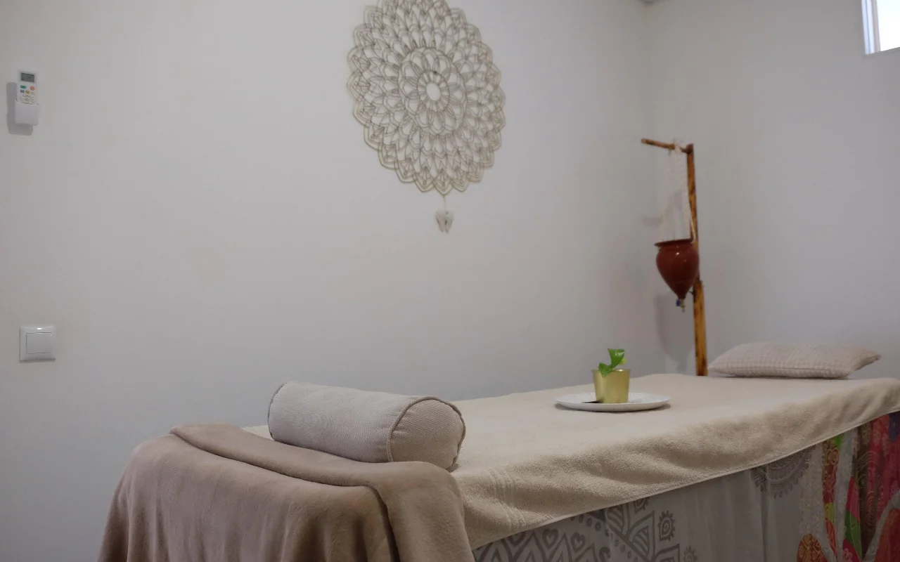 Uma sala de massagem serena em tons neutros e suaves, com uma marquesa de massagem coberta com uma toalha e um rolo de apoio, uma grande mandala em crochê pendurada na parede e um suporte de madeira que segura um vaso de terracota suspenso.