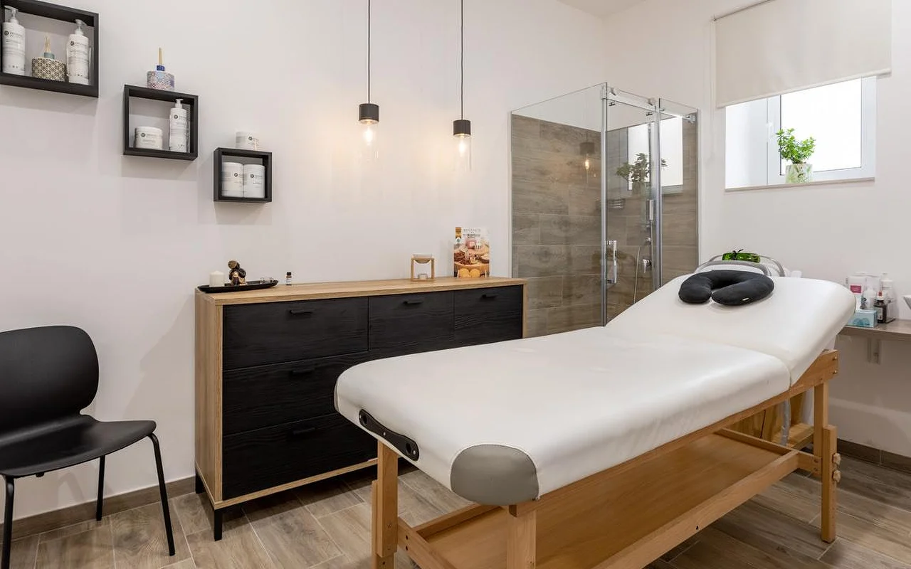 Uma sala de massagens na Thea Estetica, com uma marquesa de massagem em madeira encimada por um apoio de rosto preto, uma cómoda em preto e madeira, dois candeeiros de teto, prateleiras em cubo com produtos e uma cabine de duche em vidro com azulejo com efeito de madeira.