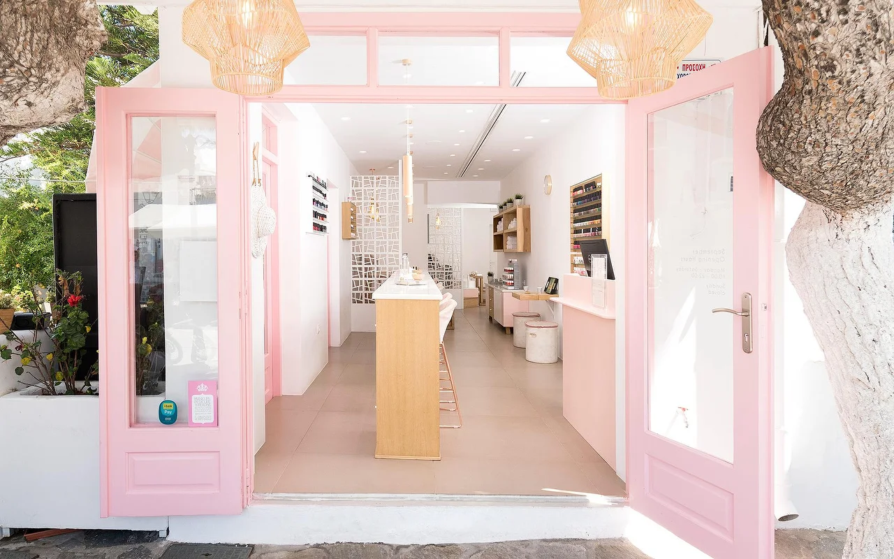 A entrada emoldurada a rosa do Vivian Nail Boutique em Naoussa, Paros, com portas abertas, candeeiros suspensos entrançados e um balcão de madeira, num edifício cicládico caiado de branco ao lado de uma árvore.