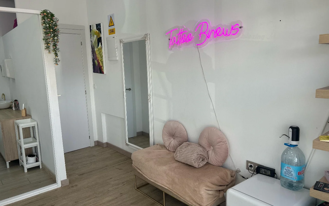 Um espaço de tratamento na YG Beauty Estudio em Torremolinos, com um letreiro néon cor-de-rosa «Tattoo Brows», um espelho de corpo inteiro e uma marquesa bege acolchoada.