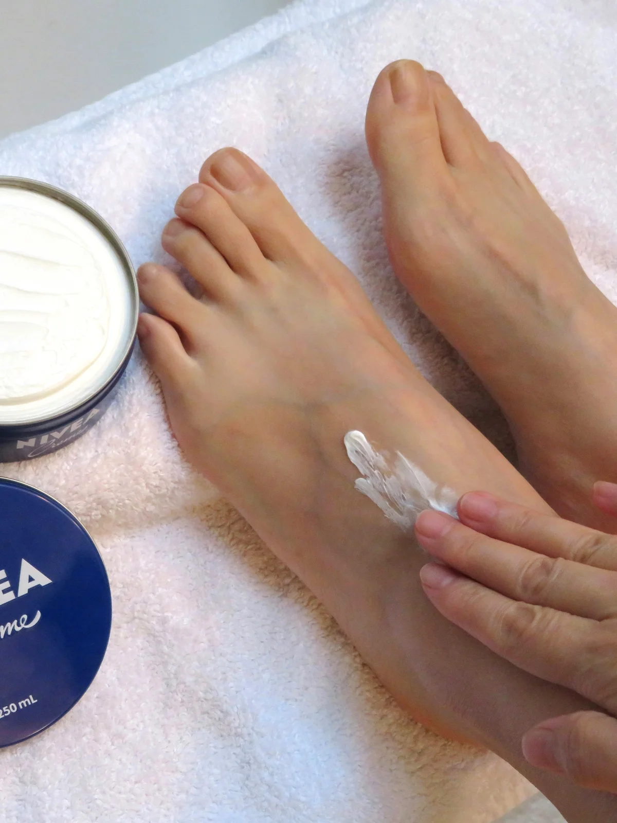 Grande plano de cima dos pés de uma mulher pousados sobre um lençol branco, com um boião de creme aberto ao lado, enquanto aplica o creme