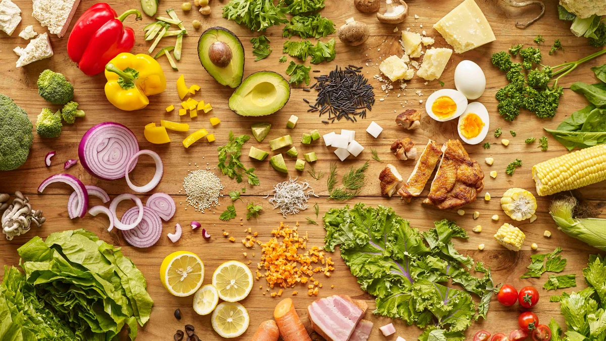 Vista de cima de uma bancada de cozinha em madeira coberta de alimentos nutritivos e coloridos: ovos, legumes, salada e frango