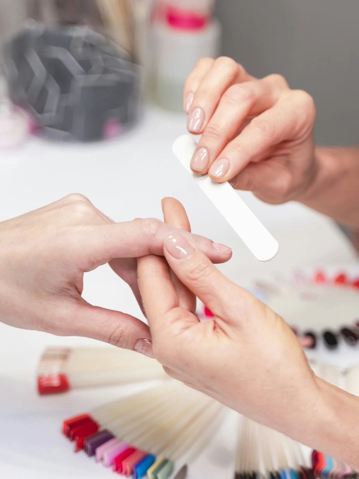 Grande plano de uma manicure profissional a limar suavemente as unhas de uma cliente no salão, com produtos e material de manicure ao fundo