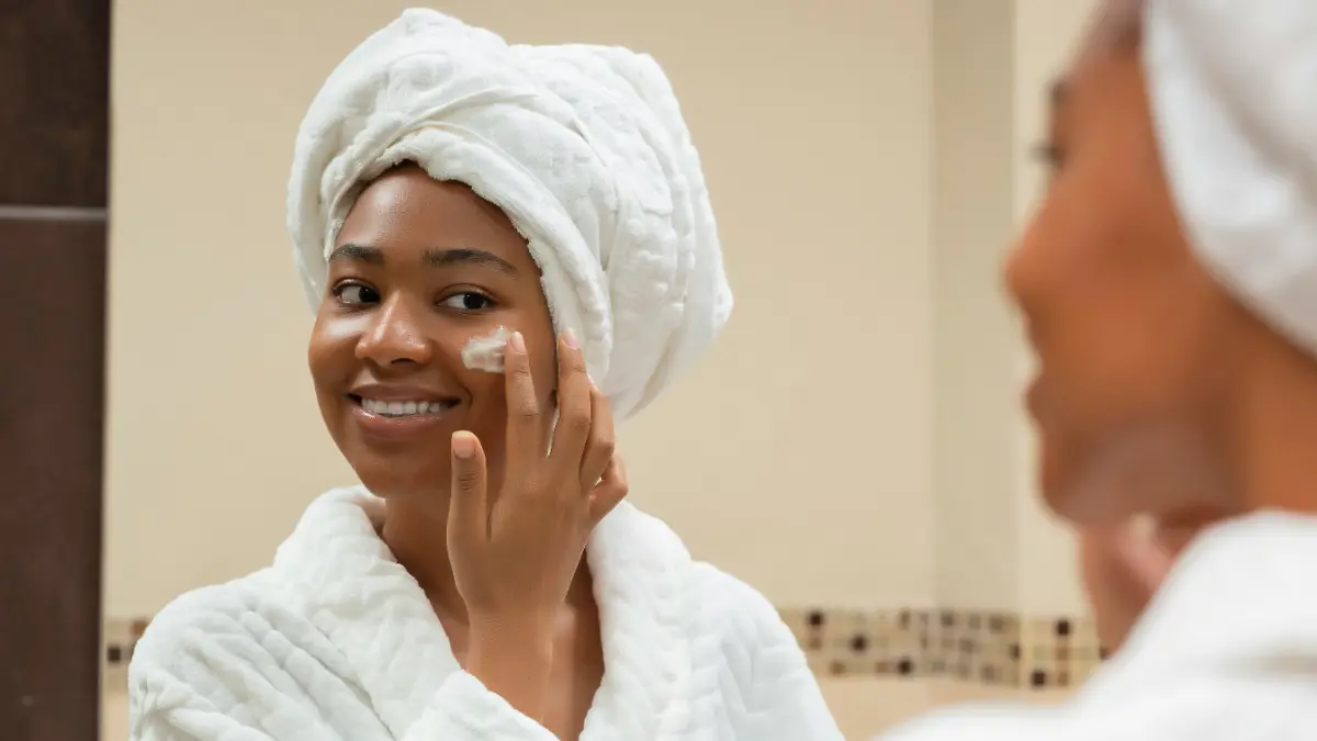 Uma mulher de robe branco com uma toalha branca enrolada na cabeça sorri ao espelho da casa de banho enquanto aplica protetor solar na cara
