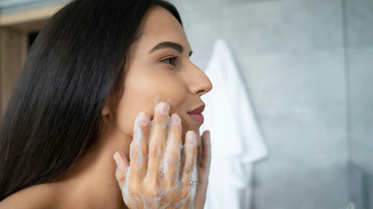 Vista de perfil de uma mulher a massajar um produto de limpeza na cara, inclinada sobre o lavatório