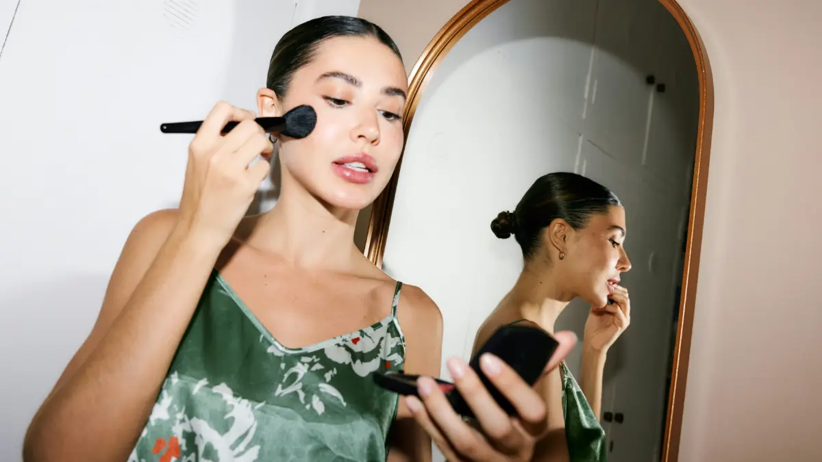 Uma mulher elegante e pronta para sair olha para um espelho de bolso enquanto aplica maquilhagem com um pincel
