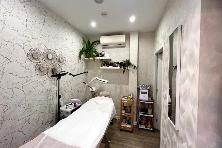 Cabine estética tranquila com uma marquesa de tratamentos coberta, uma lâmpada de aumento e um vaporizador, um carro de produtos, paredes texturadas cinzento-pálido decoradas com discos metálicos e plantas em cascata sobre as prateleiras.