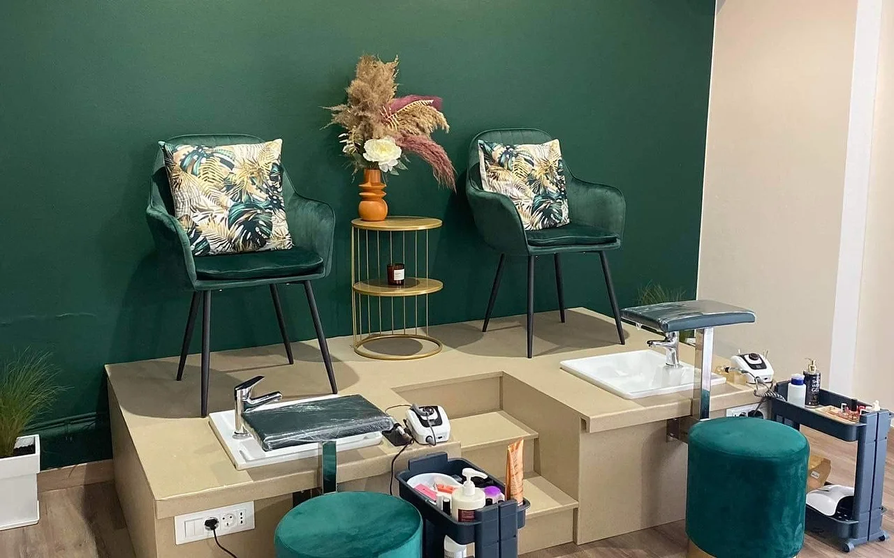 Um canto de pedicure com uma parede verde intenso, duas cadeiras tipo balde em veludo verde com almofadas de estampado tropical elevadas numa plataforma bege por cima de bacias de pedicure, uma mesinha de apoio dourada com plumas de pampa numa jarra cor de laranja, e bancos a condizer em veludo verde.