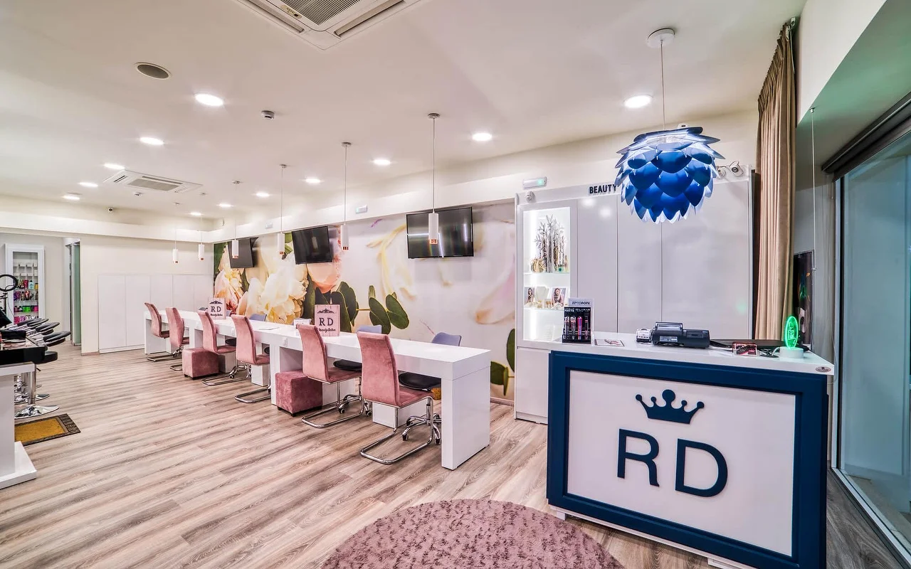 Um salão claro e branco com uma longa fila de mesas de manicure e cadeiras rosa-pálido, um mural floral suave, chão de madeira clara, e um balcão de receção marcado com um logotipo de coroa 'RD'.